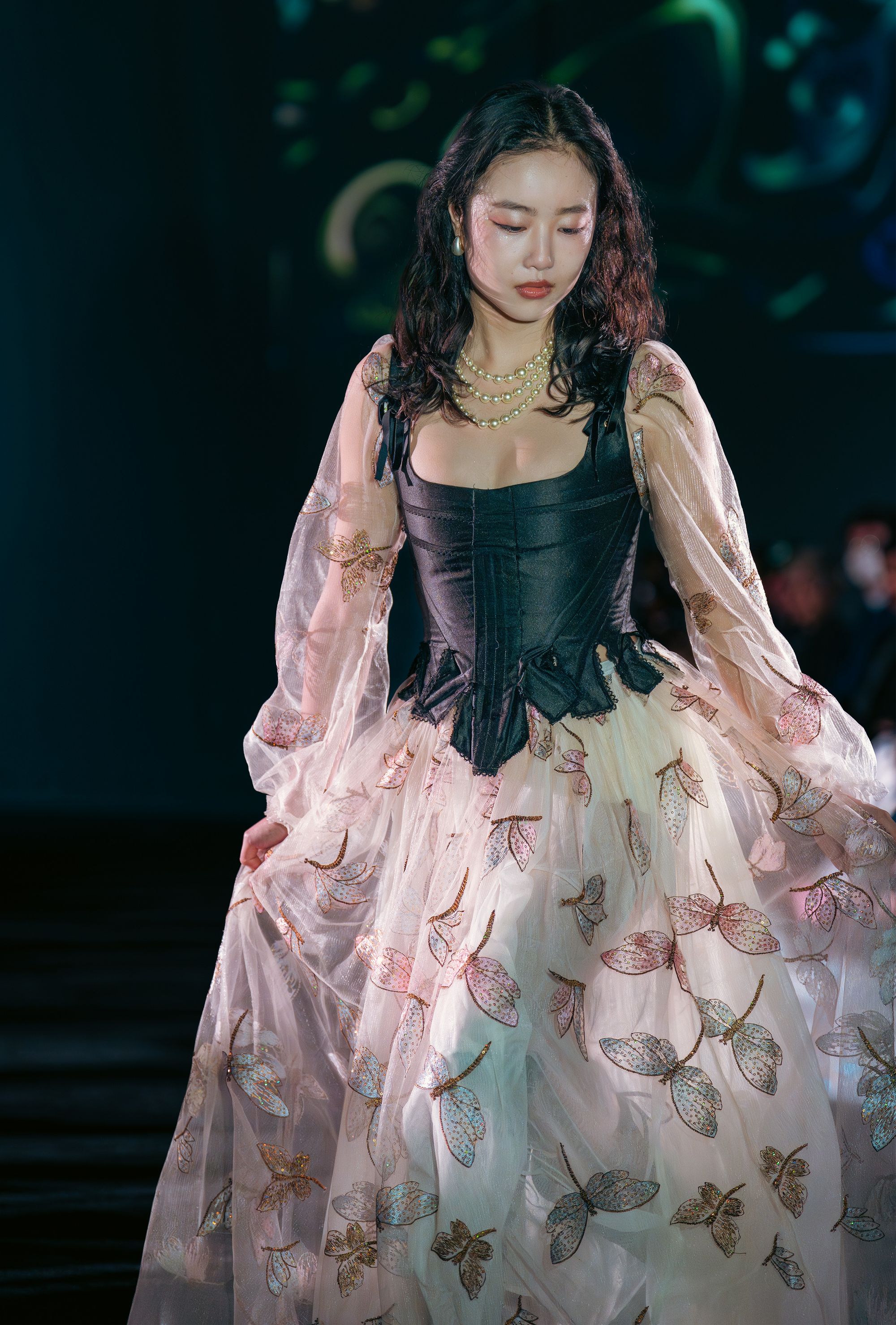 Model wears a black corset with a voluminous sheer overlay skirt embroidered with delicate multicolored butterflies. Her look is styled with soft waves and layered pearl necklaces.
